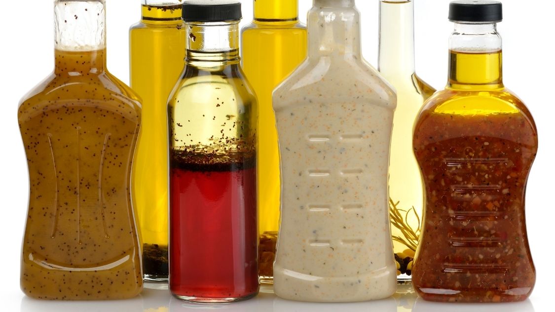 Billedet viser en samling af forskellige dressinger i gennemsigtige flasker. Fra venstre ses en krydret sennepsdressing, efterfulgt af en flaske med olivenolie. Den tredje flaske indeholder en rød vinaigrette med tydelige krydderier i bunden, og ved siden af er en cremet hvid dressing med urter og korn. Til højre ses endnu en krydret dressing med et mix af urter og krydderier synlige gennem glasset. Disse dressinger repræsenterer et bredt udvalg af smagsprofiler, der kan tilføje kompleksitet og dybde til enhver salat eller ret.