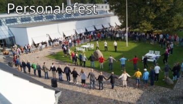 Billedet viser en stor gruppe mennesker samlet i en rundkreds til en personalefest udendørs ved en moderne bygning – en festlig og inkluderende stemning fyldt med fællesskab.