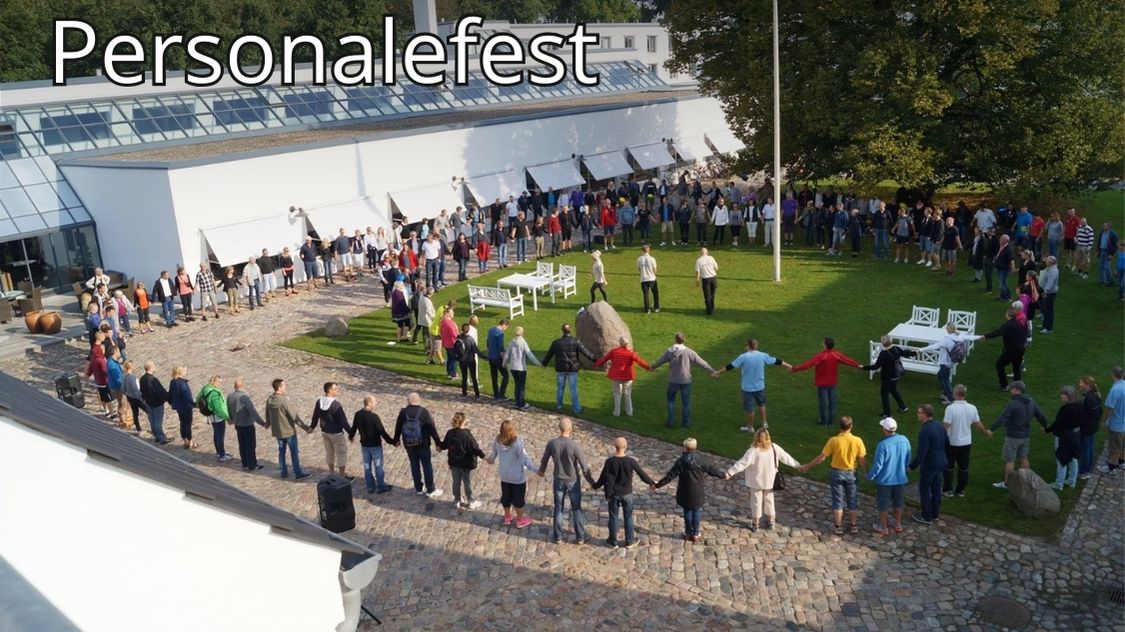 Billedet viser en stor gruppe mennesker samlet i en rundkreds til en personalefest udendørs ved en moderne bygning – en festlig og inkluderende stemning fyldt med fællesskab.