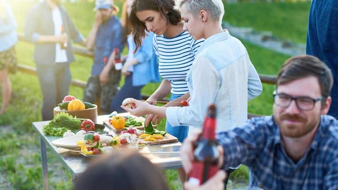 Sommerfest mad (2) Billedet viser sommerfestmad, hvor deltagerne hygger sig med at forberede friske grøntsager og grillspyd under åben himmel – en afslappet og social stemning.