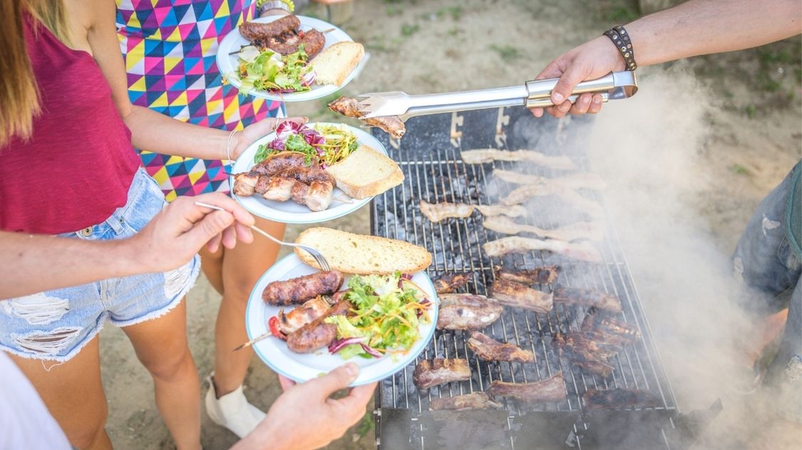 Billedet viser en livlig sommerfest med grillmad, hvor deltagerne får serveret grillede pølser, spareribs, salat og brød – en ægte uformel og hyggelig stemning.